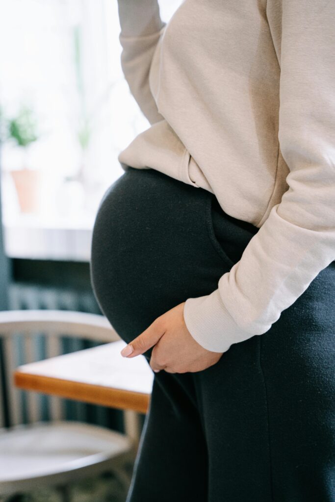 Side profile of a pregnant woman gently holding her belly wearing a white sweater and black pants.